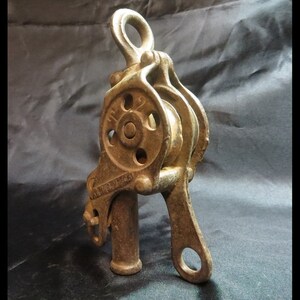 May include: A vintage cast-iron pulley system with a bronze finish. The pulley has two wheels, a top loop, and a handle with a circular opening. The text "WALKER PLIES" is visible. This antique tool is likely used for lifting or pulling, and is a classic example of engineering.