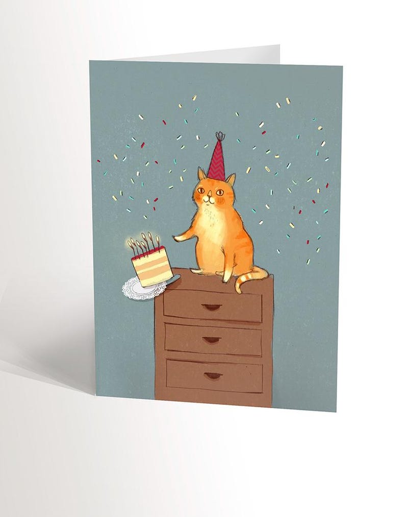 May include: A birthday card featuring an orange cat wearing a red party hat, sitting on a brown dresser with a cake with lit candles on a white doily.