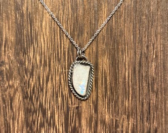 May include: A silver necklace with a pendant featuring a white, iridescent stone. The pendant has a rope-like border and is suspended from a delicate chain. The background is a wooden surface.