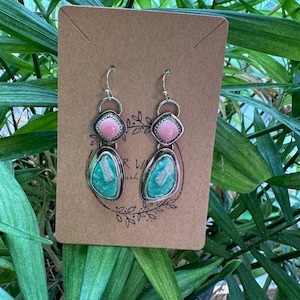 May include: A pair of handcrafted earrings with a silver metal setting. Each earring features a pink, square-shaped stone at the top and a turquoise-colored, oval-shaped stone at the bottom. The earrings are displayed on a brown card.