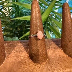 May include: A silver ring with a light pink oval gemstone. The ring is displayed on a wooden ring holder, with green foliage in the background. The gemstone has a delicate, soft color.