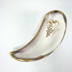 May include: A white ceramic serving dish with a gold rim and a grape design in gold. The dish is shaped like a crescent moon.