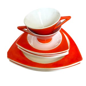 May include: A stack of triangular ceramic dinnerware featuring a vibrant orange border and a cream-colored center. The set includes a footed cup with two angular handles, matching saucers, and larger plates, all sharing the same geometric shape.