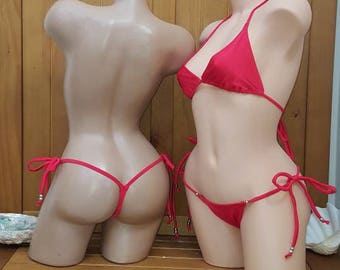 May include: Two mannequins display red bikinis. One mannequin is shown from the back, the other from the front. The bikinis have a triangle top and a thong bottom. The mannequins are on a wooden table.