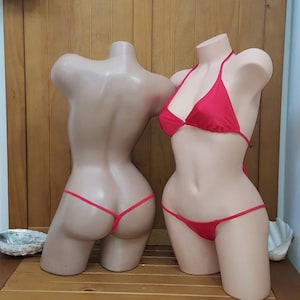 May include: Two mannequin torsos, one with a red bikini. The bikini features a triangle top and a thong bottom. The mannequins are on a wooden bench, with decorative fish and a seashell above.