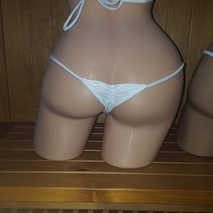 May include: White bikini bottom with a gathered front detail. The thong-style bottom is displayed on a mannequin. The string straps are tied at the sides. The mannequin is on a wooden surface.