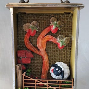 May include: A decorative box featuring a rural scene. It contains a tree with red strawberries, a brick wall, a wooden fence, and a sheep figurine. The box has a brass handle and trim, with a woven background.
