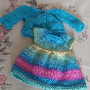 May include: A knitted baby outfit comprising a light blue cardigan with a button and a striped dress. The dress features bands of pink, blue, and yellow. The set is handmade, suggesting a soft and comfortable feel.