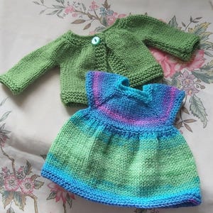 May include: A green knitted doll-sized cardigan with a single button and a blue and purple knitted dress. The dress features a gradient of colours, transitioning from blue to green and purple. The items are handmade.