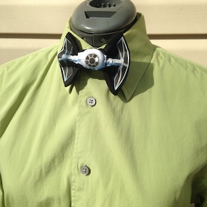 Puede incluir: Una pajarita negra con un diseño de caza TIE, con un elemento circular central blanco y azul y alas negras. La pajarita está colocada sobre una camisa verde claro con botones.