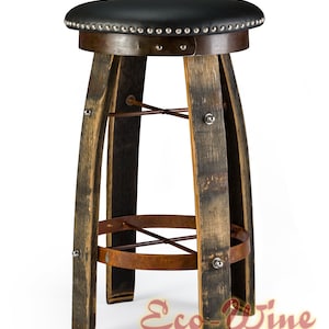 May include: Rustic wine barrel bar stool with a black leather seat featuring silver nailhead trim. The stool's legs are made from reclaimed wine barrel staves, secured with metal accents.  A rusty metal footrest adds to its industrial-chic design. Eco-Wine Furniture logo is visible at the bottom.  The overall style is vintage and repurposed.