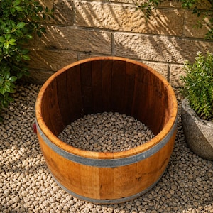 May include: A wooden barrel planter with a light brown exterior and a dark brown interior. The barrel is reinforced with metal bands and filled with small, light-colored stones. The planter sits on a bed of similar stones, with greenery in the background.