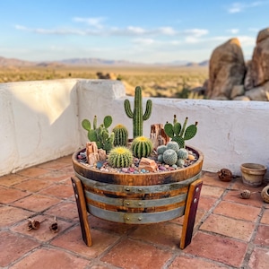 May include: A wooden barrel planter filled with various cacti and succulents, accented with colorful stones. The barrel has metal bands and wooden legs, set against a desert landscape with a blue sky.
