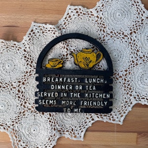 May include: Black cast iron hot pad with a teapot and teacup design. The text reads "Breakfast, Lunch, Dinner or Tea Served in the Kitchen Seems More Friendly To Me."