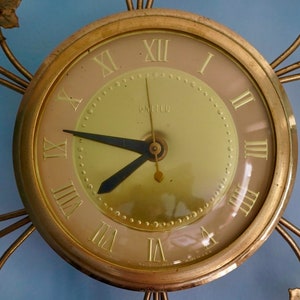 May include: A gold-toned clock with Roman numeral markings and a gold-colored metal frame with leaf accents. The clock face is cream-colored with a gold-colored center.
