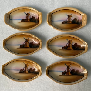 May include: Set of six small, oval-shaped ceramic dishes with a brown glaze. Each dish features a hand-painted image of a windmill with a blue and white sail against a sunset background.
