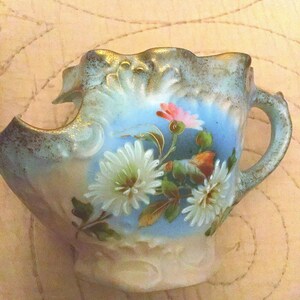 May include: A chipped, light blue porcelain teacup with a floral design. The cup has a scalloped rim and a handle. The flowers are painted in shades of white, pink, and green.