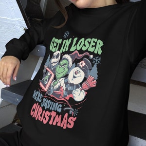 May include: Black sweatshirt featuring a graphic with the text "GET IN LOSER WE'RE SAVING CHRISTMAS." The graphic includes cartoon characters Jack Skellington, The Grinch, and a snowman. The design has a vintage, distressed look.