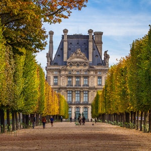 May include: A view down a tree-lined path leading to a large, ornate building with a gray roof. The trees are mostly green with some yellow leaves, and the building has a symmetrical facade with many windows.