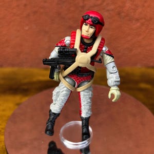 May include: A vintage action figure of a pilot in a red helmet and uniform with a black rifle. The figure has a tan harness and white and gray pants. The figure is standing on a reflective surface.