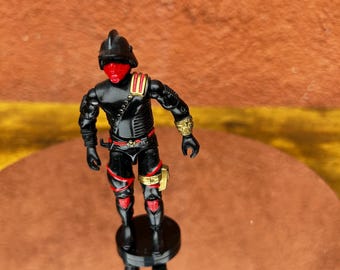 May include: A black action figure with red accents, a red face, and a black helmet. The figure has gold-colored details on the wrist and leg. The figure is standing on a black base, with its reflection visible below.