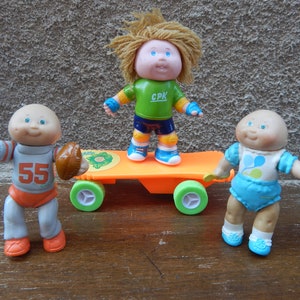 May include: Three Cabbage Patch Kids dolls, one on a skateboard, one holding a football, and one standing with a blue and yellow shirt. The dolls have blonde hair and blue eyes.
