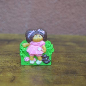 May include: A plastic figurine of a Cabbage Patch Kid doll with brown hair, wearing a pink dress and holding a black cat. The doll is standing in a green plastic base with flowers.