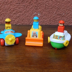 May include: Three toy vehicles with figures on top. A blue airplane with a yellow propeller, a yellow bulldozer with an orange blade, and a green and white boat with a yellow propeller.