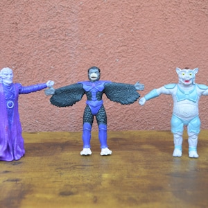 May include: Three action figures, one purple, one blue and one black and blue, standing on a wooden surface.