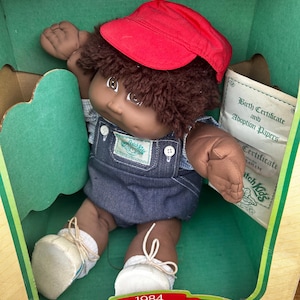 May include: A vintage Cabbage Patch Kids doll in its original box. The doll has brown skin, brown hair, and is wearing a red cap, denim overalls, and white shoes. The box includes a birth certificate and adoption papers. The box reads "1984 Cabbage Patch Kids".