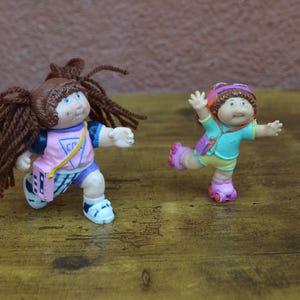May include: Two Cabbage Patch Kids dolls, one with brown hair and a pink and blue outfit, the other with brown hair and a blue and green outfit, both are wearing roller skates.