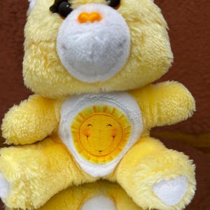 May include: A yellow plush bear with a white muzzle and belly patch. The bear has black eyes and an orange nose. The belly patch features a smiling sun design. The bear is a children's toy.