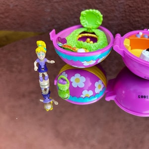 May include: A pink and yellow plastic egg-shaped toy with a green lid. The egg is decorated with white flowers. Inside the egg is a small green playset with a yellow chick. A small blonde doll with a purple dress is standing next to the egg.