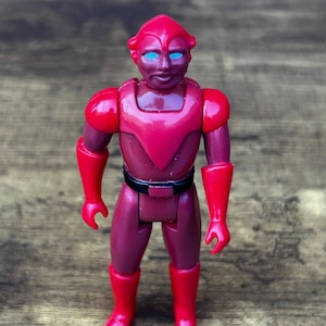 May include: A vintage action figure of a humanoid alien in shades of red. The figure has a red helmet, shoulder pads, gloves, and boots. It has a black belt and blue eyes. The figure is standing on a wooden surface.