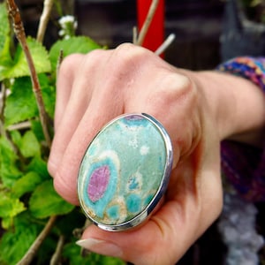 May include: A silver ring featuring a large oval green and pink gemstone. The gemstone has a swirling pattern of green, pink, and white.