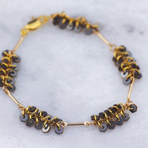 May include: A gold-toned bracelet with a delicate design. The bracelet features small, dark gray hexagonal beads linked together with gold-colored metal. The clasp is also gold-toned. The bracelet is displayed on a white surface.