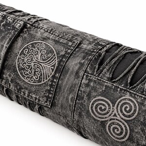 May include: A cylindrical bolster pillow featuring a charcoal gray acid-wash fabric finish. The design includes a circular embroidered tree motif and a triple spiral triskelion symbol, accented by decorative black fabric lacing along the sides.