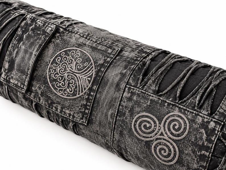 May include: A cylindrical bolster pillow featuring a charcoal gray acid-wash fabric finish. The design includes a circular embroidered tree motif and a triple spiral triskelion symbol, accented by decorative black fabric lacing along the sides.
