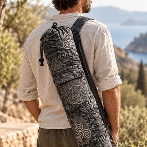 May include: A cylindrical black acid-wash denim yoga mat bag featuring a drawstring closure at the top, a shoulder strap, and white embroidered circular motifs including a central tree of life design flanked by two triskelion symbols.