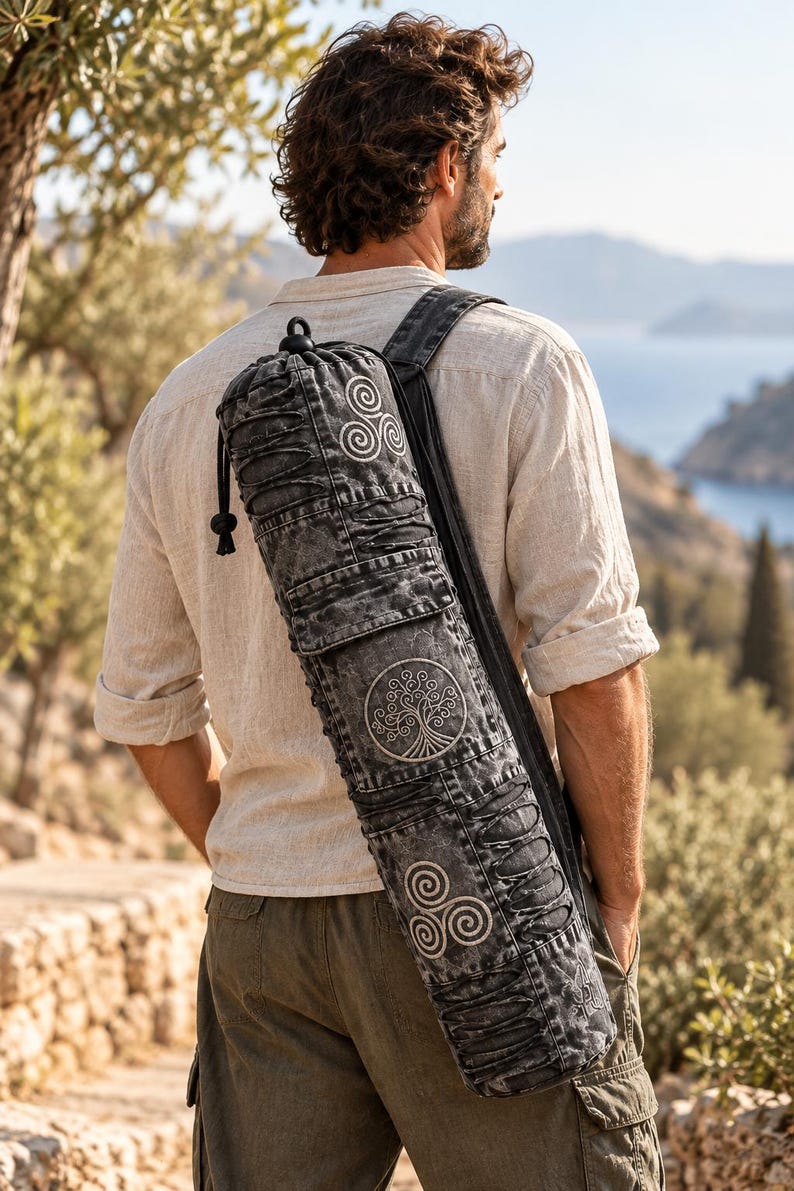 May include: A cylindrical black acid-wash denim yoga mat bag featuring a drawstring closure at the top, a shoulder strap, and white embroidered circular motifs including a central tree of life design flanked by two triskelion symbols.