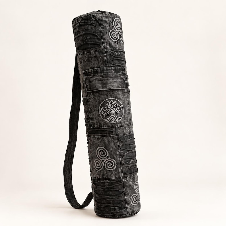 May include: A cylindrical yoga mat bag in a dark gray acid-wash fabric. The bag features embroidered light gray circular symbols, including a tree of life and triskelion designs, along with horizontal stitched cut-out details and a shoulder strap.