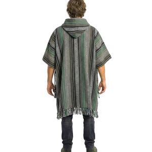 May include: A hooded poncho featuring a vertical striped pattern in shades of forest green, black, white, and grey. The garment has short sleeves, a hood, and a fringed hemline at the bottom. It is worn over dark grey jeans and dark shoes.