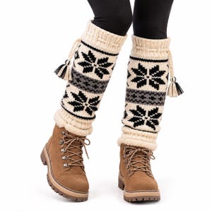 May include: A pair of cream-colored knit leg warmers featuring a black and gray snowflake pattern, ribbed cuffs, and decorative braided drawstrings with black and cream tassels, worn over brown lace-up boots with thick lug soles and black leggings.