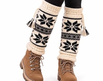 May include: A pair of cream-colored knit leg warmers featuring a black and gray snowflake pattern, ribbed cuffs, and decorative braided drawstrings with black and cream tassels, worn over brown lace-up boots with thick lug soles and black leggings.