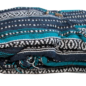 May include: A folded woven blanket featuring a dark navy blue, bright teal, and white geometric pattern. The textile displays horizontal stripes with intricate diamond and dash motifs across the surface, with visible frayed threads at the bottom edge.