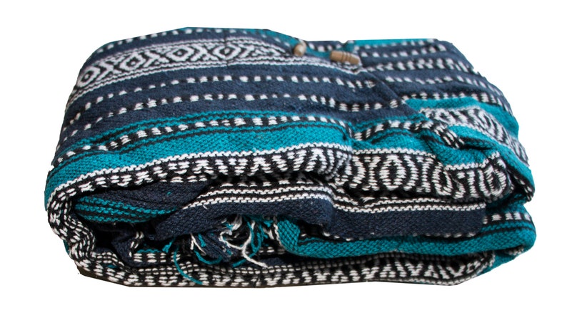 May include: A folded woven blanket featuring a dark navy blue, bright teal, and white geometric pattern. The textile displays horizontal stripes with intricate diamond and dash motifs across the surface, with visible frayed threads at the bottom edge.