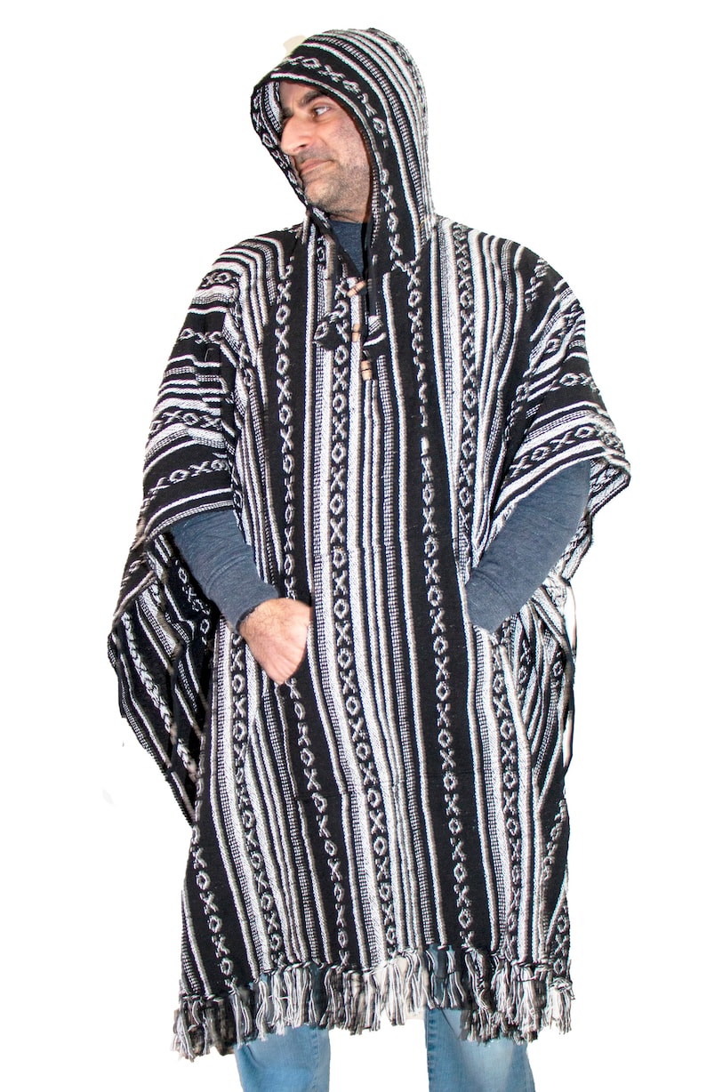 May include: A black and white hooded poncho featuring a woven pattern of vertical stripes and repeating X-shaped motifs. The garment includes a front kangaroo pocket, a drawstring hood with wooden beads, and a fringed hem along the bottom edge.