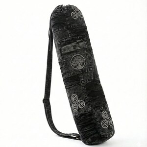 May include: A cylindrical yoga mat bag in a black acid-wash denim fabric. The bag features a drawstring closure at the top, an adjustable shoulder strap, and embroidered light gray circular symbols including a tree motif and triskelion designs.