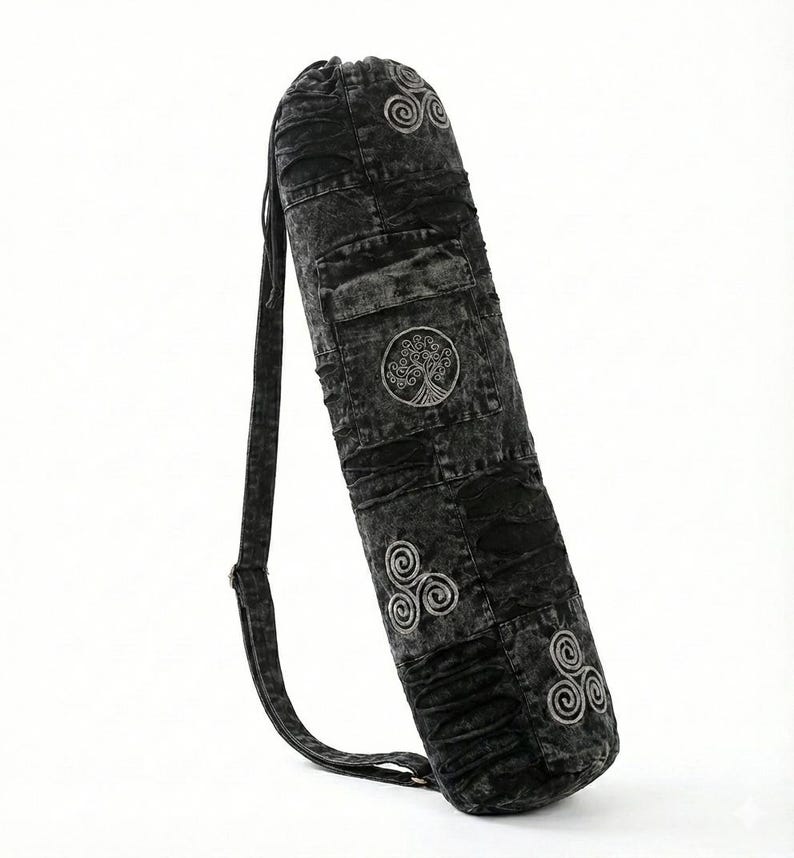 May include: A cylindrical yoga mat bag in a black acid-wash denim fabric. The bag features a drawstring closure at the top, an adjustable shoulder strap, and embroidered light gray circular symbols including a tree motif and triskelion designs.