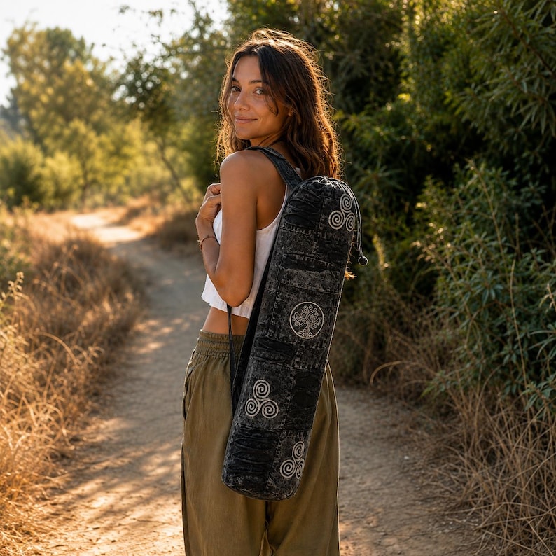 May include: A black, acid-washed cylindrical yoga mat bag featuring white embroidered symbols, including a central tree of life design and multiple triskelion patterns, with a drawstring closure and a single adjustable shoulder strap.